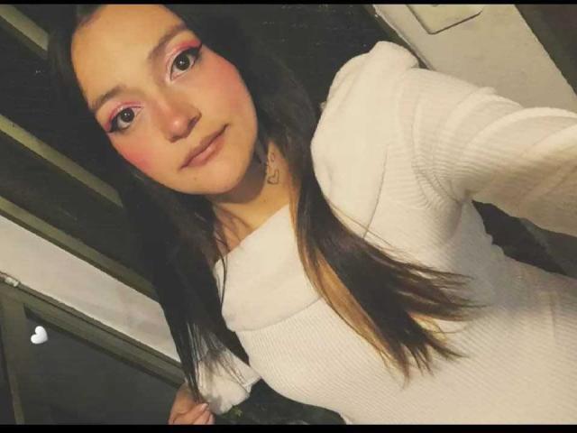 MollyWilson has long, straight brown hair, fair skin, and pink eye makeup with dark eyeliner. She's wearing a white, ribbed, long-sleeve top with a cowl neck. Her expression is neutral with slightly parted lips. The lighting is soft, highlighting her pink blush on her cheeks. The image has a slight digital filter, giving it a warm, slightly grainy texture. She's holding the camera with her right hand.