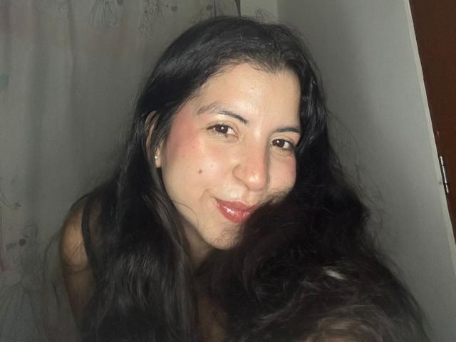 Close-up of IanaPrince with long, wavy black hair, fair skin, and brown eyes. She has a small mole on her right cheek and a subtle smile. Her pink lips are slightly parted. She's wearing small pearl earrings. The light source is from the front, casting soft shadows. Her hair partially covers her right cheek. The image has a natural, candid feel.