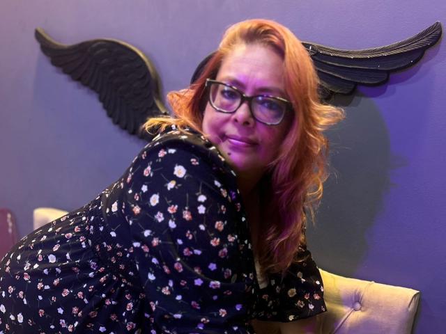 MiluSmith has red-orange hair, glasses, and is smiling while leaning on a couch, wearing a black floral dress. Black angel wings are in the background.
