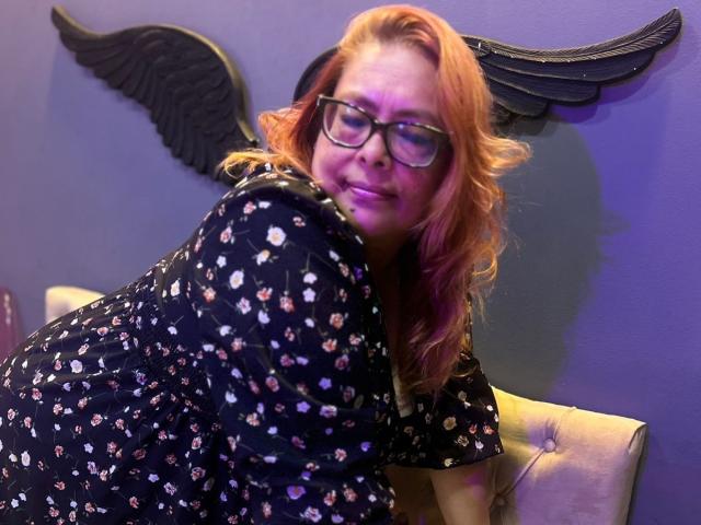 MiluSmith with wavy orange hair and glasses, smiling while leaning on a tufted chair, wearing a black floral dress, her cute ass slightly visible.
