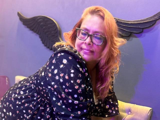 MiluSmith with wavy orange hair, glasses, and black floral dress, leaning forward, smiling, butt facing camera, blue wall with black wing decoration in background.