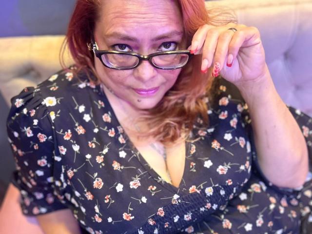 MiluSmith with glasses, red nails, and pink lipstick, wearing a floral black dress, showing her tits, smiling, and holding her glasses. Her red hair is slightly wavy.