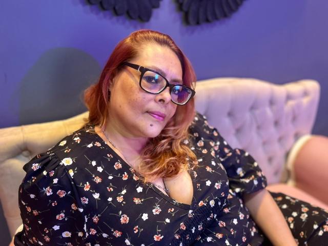 MiluSmith with red hair, glasses, and a black floral dress, sitting on a beige tufted sofa, showing her boobs in a slightly low-cut dress. She's looking at the camera.
