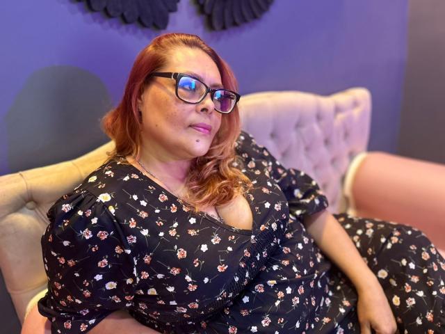 MiluSmith, a plus-size woman with red hair, wears black floral dress with puffy sleeves, sitting in a beige tufted chair. She has glasses, a small mole on her cheek, and a confident, relaxed expression. The background is a purple wall with black decorative elements. The lighting is warm, highlighting her fair skin and the texture of her dress. Her hands rest on her thighs, and her right arm is slightly raised.