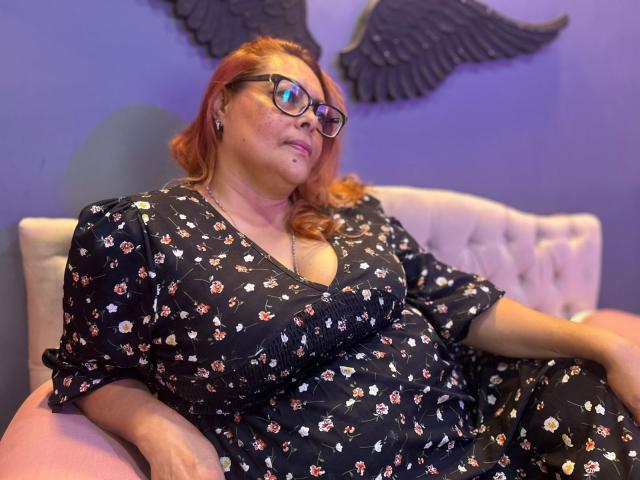 MiluSmith, a chubby woman with red hair and glasses, is sitting on a pink tufted couch wearing a black floral dress, showing her tits and a small nose piercing.