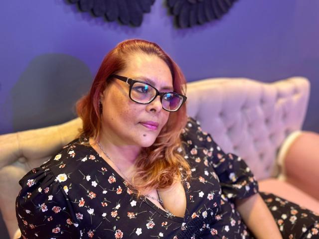 MiluSmith with glasses, red hair, and freckles, wearing a black floral dress, sitting on a pink couch, showing her cleavage. Her pink lips and slight smile are visible.