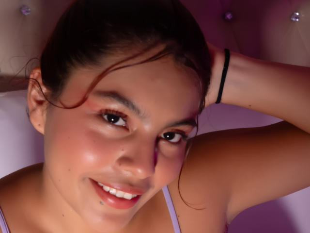AlyssaGreen's selfie shows her smiling with a lip piercing, brown eyes, and a black hair tie on her raised arm. She's wearing a pink top, with pink-tinted lighting.