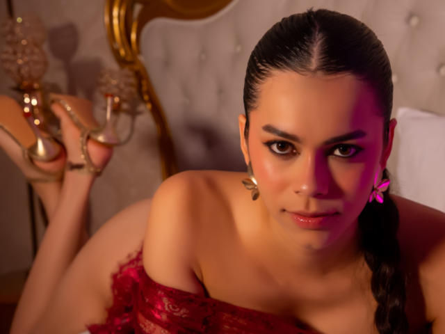 AlanaBlake with dark hair in a braid, pink bow, red lace top, and gold heels, lying on a bed, looking at the camera with a slight smile.