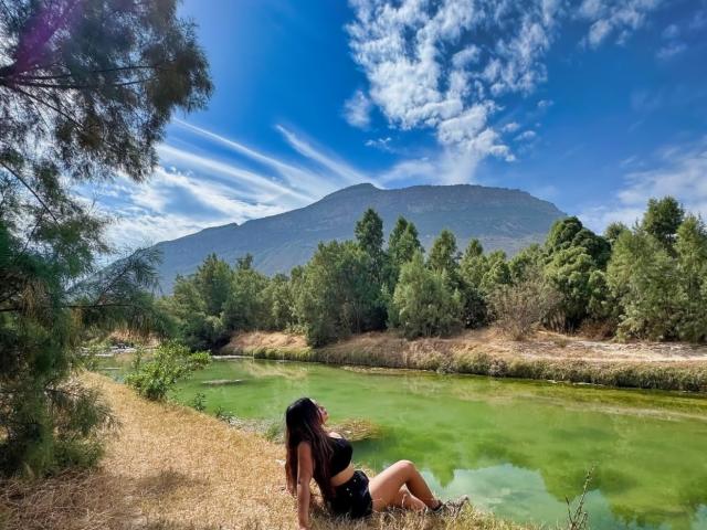 KiaraXMoon, sitting on grassy bank, back to camera, black top, shorts, green-tinted water, forest, mountain, clear blue sky, white clouds, pine trees, summer, legs up, relaxed.