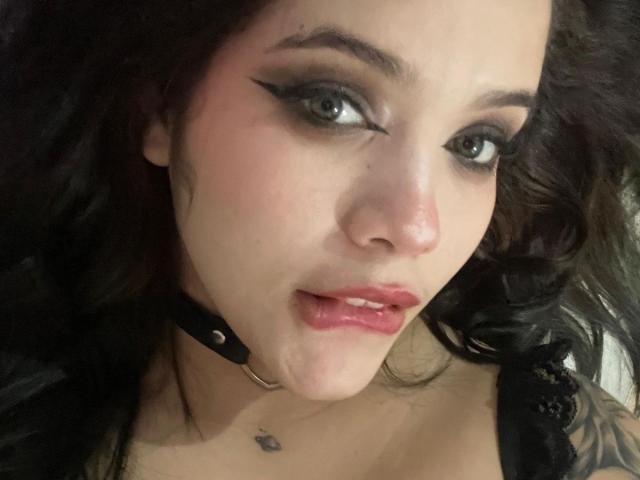 AryButty has dark eyeliner, pale skin, and a small nose piercing. Her pink lips are slightly parted. She wears a black choker with a silver ring.