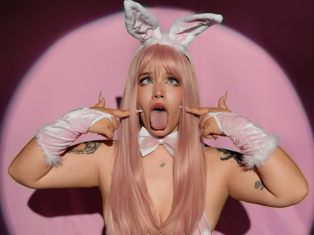 AryButty with pink hair, white bunny ears, and gloves, sticking out her tongue, pointing fingers near her mouth, tattoos on her shoulders, and a pink bowtie.