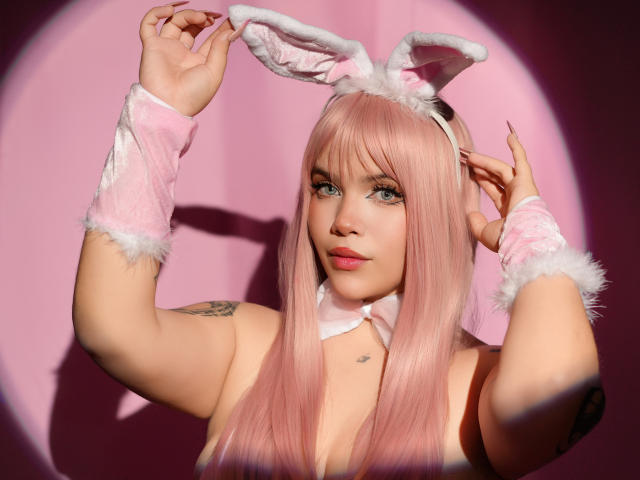 AryButty with pink hair, white furry bunny ears, pink wrist cuffs, and a pink collar, raising hands, showing a tattoo on her right arm, and a small heart tattoo.