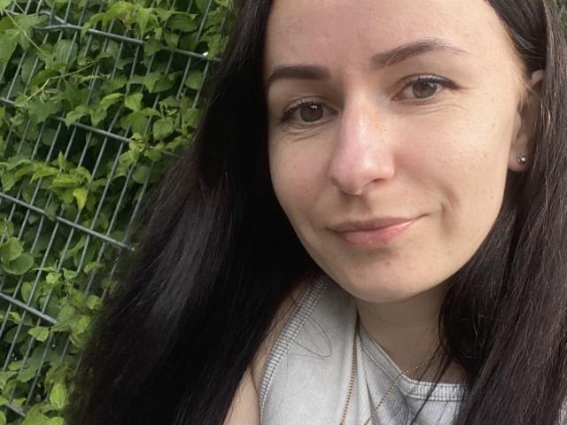 Close-up photo of DreamyAnita with light skin and long dark hair. She has brown eyes, thin eyebrows, and a small smile. Her nose is slightly upturned, and she has small earrings. She's wearing a sleeveless gray top. The background has green leafy vines and a wire fence. The lighting is natural, and the image is focused on her face. Her expression is calm and relaxed. The texture of her hair is smooth, and her skin is clear.