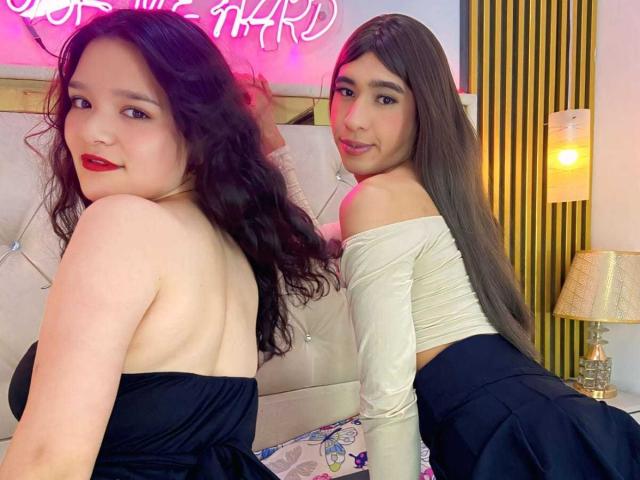 Due ragazze latine con lunghi capelli castani, rossetto rosso e abiti neri/bianchi, che mostrano la schiena e i glutei scoperti, sedute su un letto, che guardano oltre le spalle e sorridono..