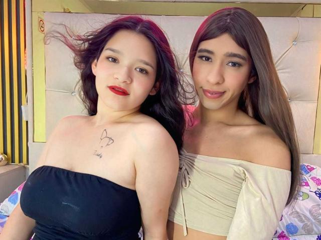 Deux femmes d'origine latine, avec de longs cheveux, dont une avec des mèches roses, portant des hauts à épaules dénudées, assises sur un lit. L'une a un haut noir, de petites seins, et un tatouage ; l'autre a un haut beige, et n'a pas de seins..