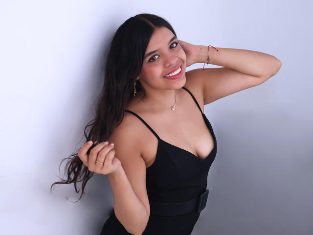 LiinaSantacruz with long black hair, smiling, wearing a black dress with a low neckline, showing her small boobs, standing against a white wall, hand in hair.