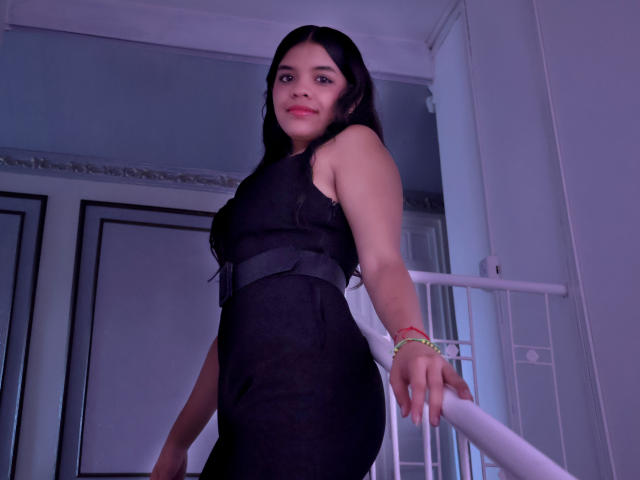 LiinaSantacruz with long black hair, wearing a black dress with a belt, standing on stairs, looking down, hand on railing, purple lighting, red bracelet, slightly smiling.