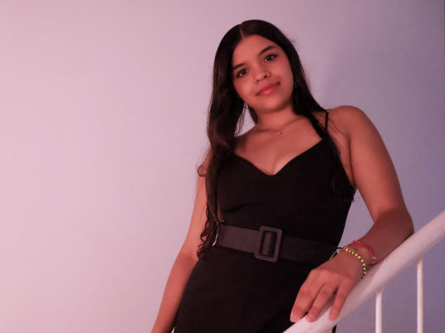 LiinaSantacruz with long dark hair, wearing a black dress with a gray belt, standing and leaning on a white railing, smiling slightly, brown eyes, and a green bracelet.