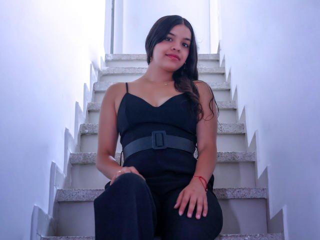LiinaSantacruz, sitting on white stairs, wearing a black dress with a belt, has long dark hair, a cute face, small tits, and a red bracelet, looking down.