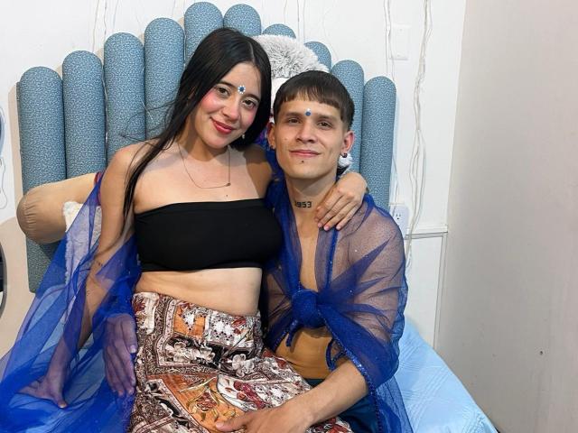 AnjaliRohan with long black hair, medium tits, and a blue sheer robe, sits next to a short-haired man with a blue robe and patterned pants, both smiling.