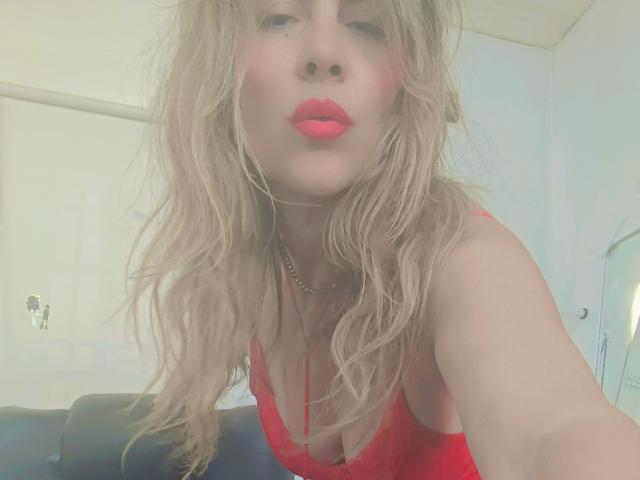 Maytena with wavy blonde hair, red lipstick, and red dress, leaning forward, showing cleavage, wearing a chain necklace, taking a selfie, looking at the camera.