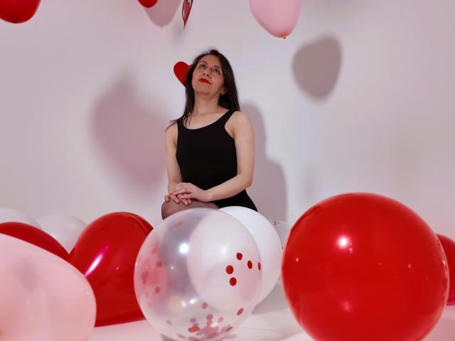 Cyntiia with long black hair, wearing a black tank top, sits among red and white balloons, hands on her knees, looking up with a slightly surprised expression.