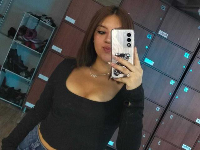 ZoeyClark with brown hair, black long-sleeve top, showing her tits in a mirror selfie, holding a white phone with a marble case, wearing a necklace, in a locker room.