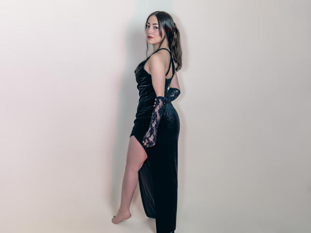 AnnFisher in a black velvet dress with lace sleeves, showing her ass with a high slit, standing barefoot, looking over her shoulder, dark hair, red lipstick.