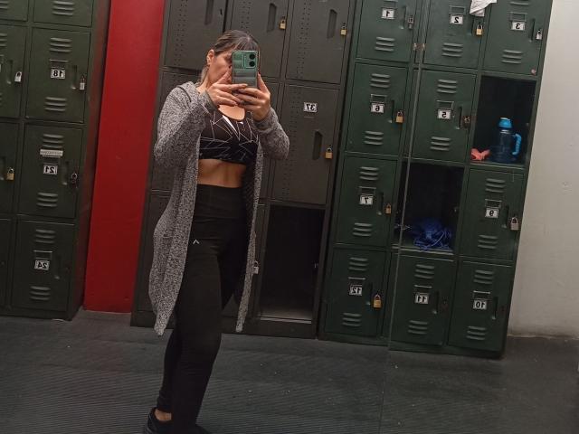 SophySheikh taking a mirror selfie in a gym locker room, wearing a gray cardigan, black sports bra, and black pants, showing her toned abs and small tits.