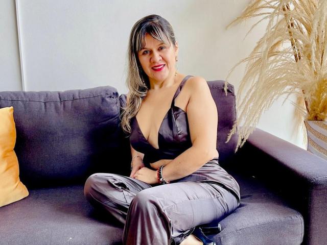 SophySheikh with gray hair, red lipstick, and black satin dress, sitting on gray couch, legs crossed, showing her tits in deep V, smiling, yellow pillow on left.