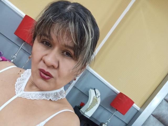 SophySheikh with short, blonde-streaked hair, wearing white lace choker and strap, red lipstick, looking at camera, small earrings, yellow and gray background, red lamps.