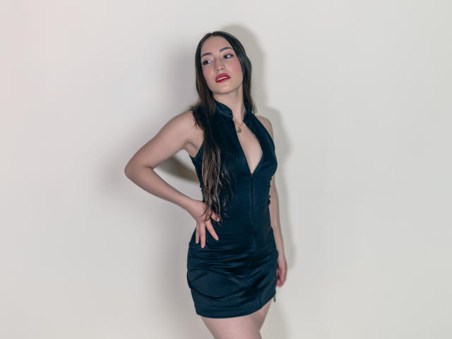 AnnFisher with long brown hair, red lipstick, wearing a black zip-up dress, showing her tits, standing with one hand on her hip, looking to the side, pale skin.