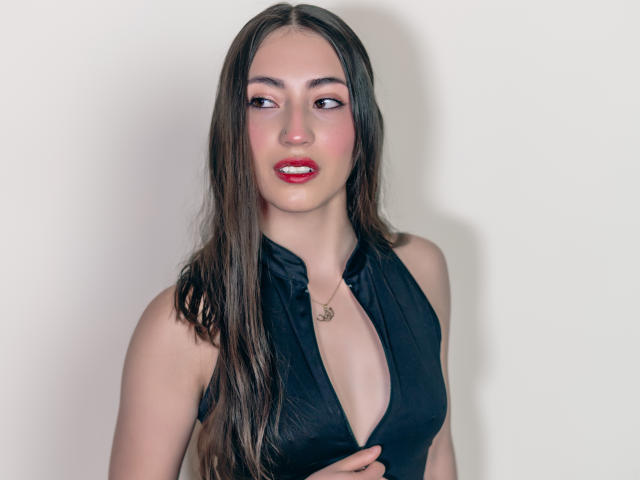 AnnFisher with long brown hair, red lipstick, and black halter top showing her tits, holding her top open, wearing a small necklace, looking to the side, smiling slightly.