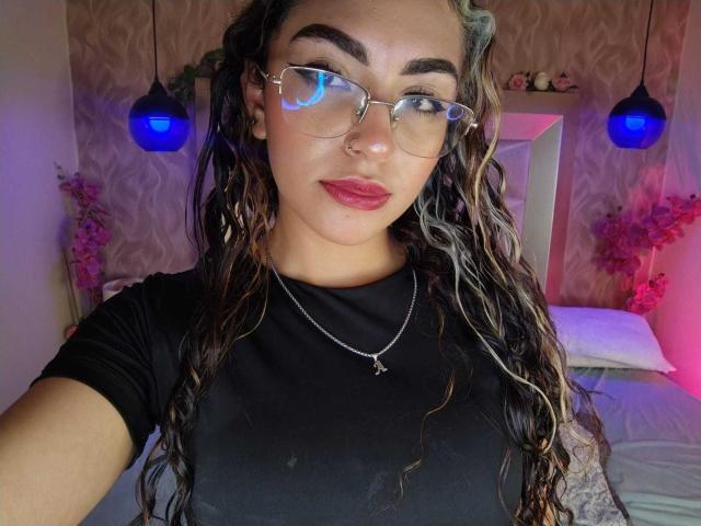 AdaraAngel with wavy brown hair, glasses, nose ring, red lipstick, black shirt, necklace, medium tits, and a confident smile, standing in a room with blue lights.