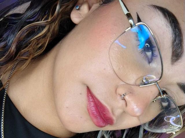 AdaraAngel with glasses, nose ring, red lipstick, wavy brown hair with blonde highlights, and a slight blush on her cheeks, looking down with a serious expression.