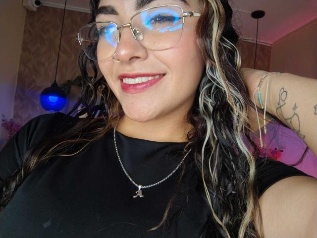 AdaraAngel with wavy black and blonde hair, glasses reflecting blue light, black shirt, red lipstick, nose ring, tattoo on right arm, smiling, necklace with cross, hand behind head.