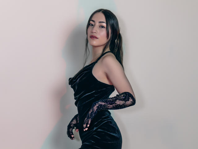 AnnFisher wearing a black velvet dress with lace gloves, standing sideways, showing her ass, long dark hair, and a confident expression. She's looking at the camera.