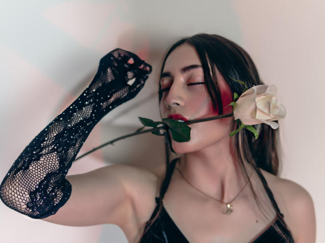 AnnFisher with eyes closed, black lace glove, holding a rose between her red-painted lips, wearing a black top, gold necklace, and pale skin. She's got a rose in her mouth.