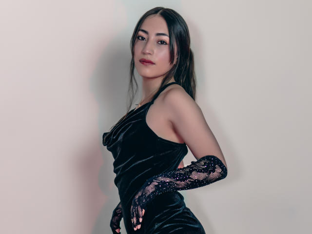 AnnFisher with long black hair, wearing a black velvet dress and lace gloves, looking at the camera with a slight smile, standing against a plain wall.