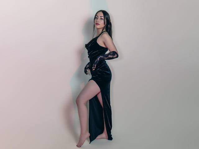 AnnFisher in a black velvet dress with a thigh-high slit, lace gloves, and bare feet, standing against a wall, looking over her shoulder with a slight smile.