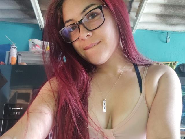 Thalassa with red hair, glasses, and a small smile, wearing a pink tank top showing her tits, necklace with dog tag, in a room with blue walls.