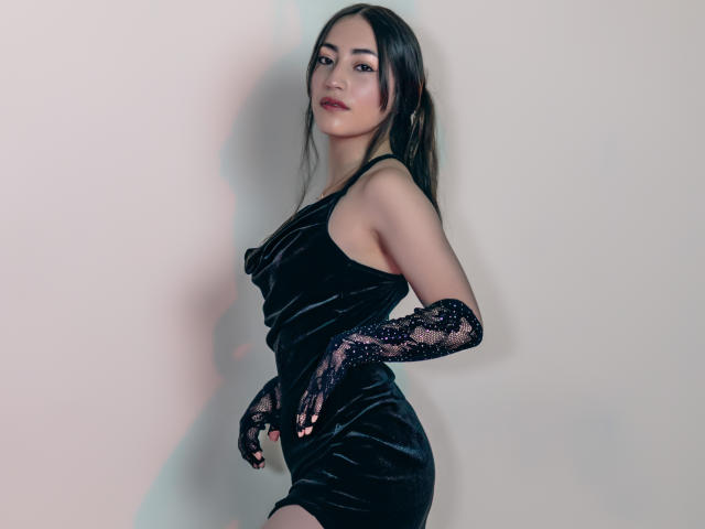 AnnFisher with long dark hair, wearing a black velvet dress and lace gloves, standing sideways, looking at camera, showing her ass, with a slight smile, in a studio.