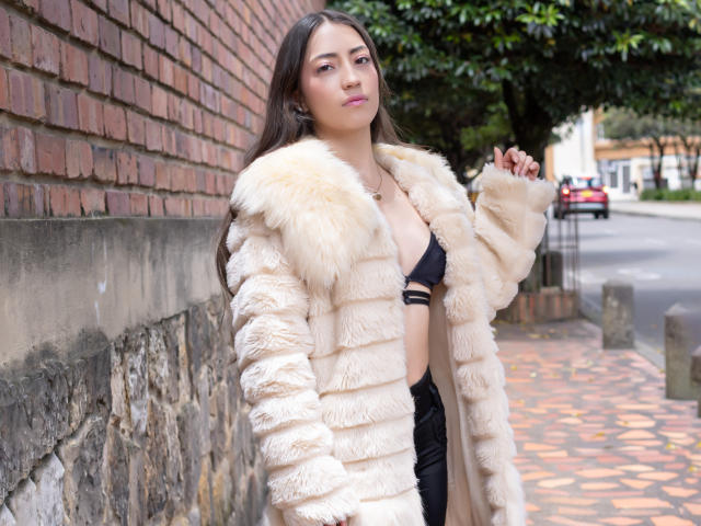 AnnFisher with long brown hair, wearing a fluffy beige fur coat, exposing black bra, standing against brick wall, looking slightly annoyed, in a street setting, hands up.