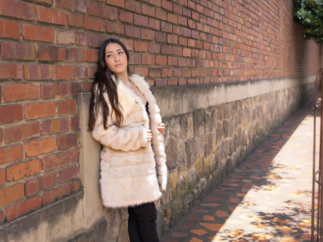 AnnFisher leaning against a brick wall in a fluffy beige fur coat, black pants, and long brown hair. Her hand rests on her coat, looking slightly to the side.