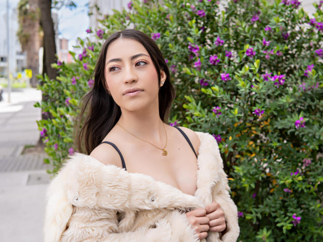 AnnFisher with dark hair, wearing a fluffy beige coat, black strap dress, gold necklace, standing in front of purple flowers, looking up with a slight smile, city background.