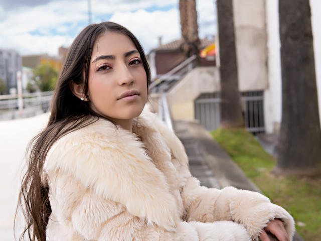 AnnFisher with long brown hair, wearing a fluffy beige coat, looking at the camera, soft pink makeup, small earrings, standing outside, overcast sky, slightly blurred background.