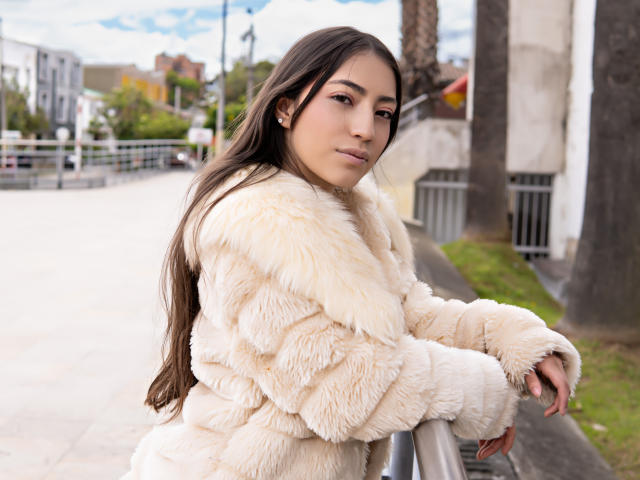 AnnFisher with long brown hair, wearing a fluffy beige fur coat, leaning on a metal railing, looking to the side with a slight smile. Her pussy is not visible.