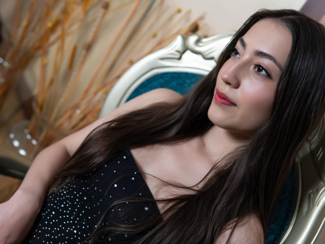 AnnFisher with long brown hair, wearing a black sparkly dress, sitting on a blue upholstered chair, smiling softly, pink lipstick, fair skin, brown eyes, looking to the side.