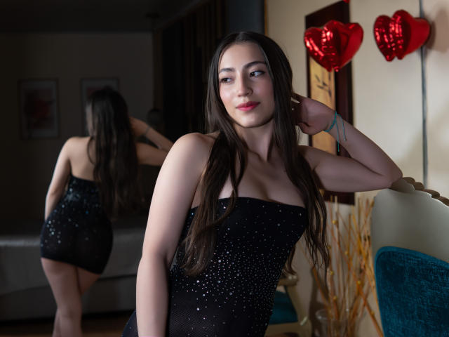 AnnFisher with long brown hair, wearing a black sparkly dress, showing her tits, standing in front of a mirror, red heart balloons in the background, smiling at the camera.