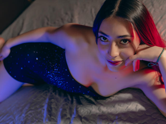 AnnFisher with red highlights, lying on a bed in a dark blue sparkly dress, her hand touching her cheek, blue and red lights on her face, showing her tits.